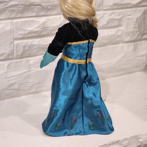 Peut inclure: Poup&eacute;e blonde coiff&eacute;e en tresse, v&ecirc;tue d'une robe en satin bleu turquoise avec des bordures dor&eacute;es et des broderies florales. Le corsage de la poup&eacute;e est noir, avec des gants turquoise.