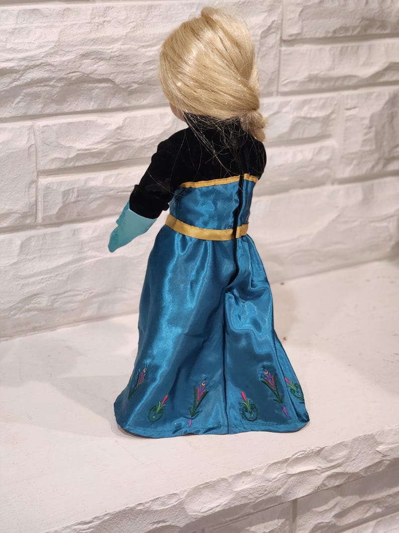 Peut inclure: Poup&eacute;e blonde coiff&eacute;e en tresse, v&ecirc;tue d'une robe en satin bleu turquoise avec des bordures dor&eacute;es et des broderies florales. Le corsage de la poup&eacute;e est noir, avec des gants turquoise.