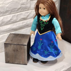 Pu&ograve; includere: Bambola con lunghi capelli castani, indossa un vestito blu con ricami floreali e un gilet nero con dettagli verdi e rosa. La bambola ha gli occhi verdi ed &egrave; in piedi accanto a una piccola scatola di legno.