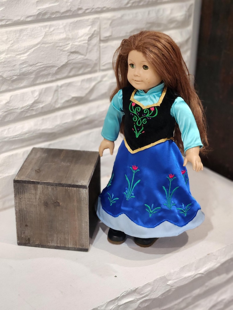 Pu&ograve; includere: Bambola con lunghi capelli castani, indossa un vestito blu con ricami floreali e un gilet nero con dettagli verdi e rosa. La bambola ha gli occhi verdi ed &egrave; in piedi accanto a una piccola scatola di legno.