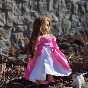 May include: A doll with long brown hair wearing a pink and white princess dress. The dress has a pink bodice and sleeves, with a white skirt. The doll is standing outdoors near a stone wall.