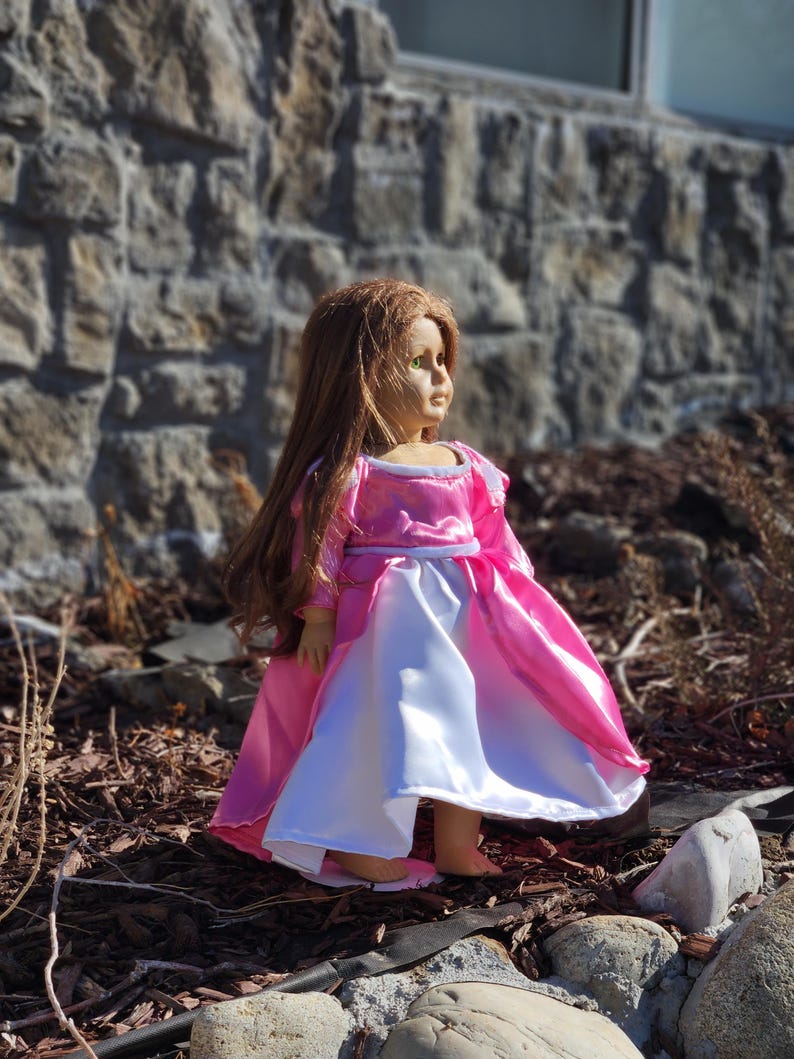 May include: A doll with long brown hair wearing a pink and white princess dress. The dress has a pink bodice and sleeves, with a white skirt. The doll is standing outdoors near a stone wall.
