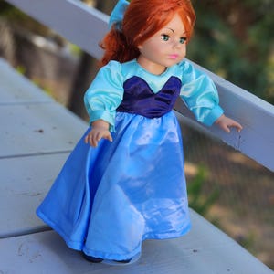 May include: A doll with vibrant red hair styled with a blue bow, featuring green eyes and pink lips. The doll wears a dress with a dark purple velvet bodice, light blue satin puff sleeves, and a matching light blue satin skirt that reaches the floor.
