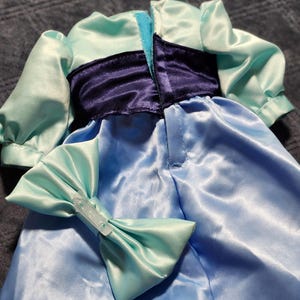 May include: A light blue satin dress featuring short puff sleeves, a dark purple waistband, and a matching light blue bow accessory. The garment has a vertical opening at the center back with a hook and loop closure, and the fabric has a smooth, reflective finish.
