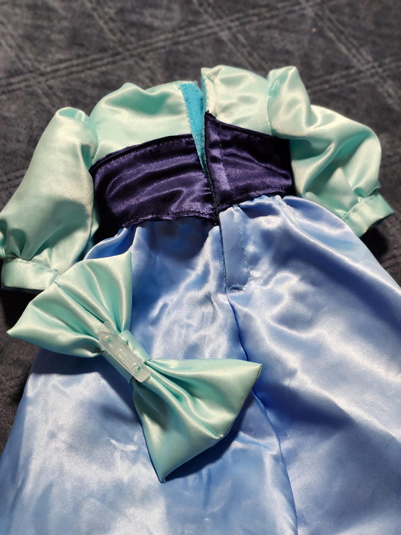 May include: A light blue satin dress featuring short puff sleeves, a dark purple waistband, and a matching light blue bow accessory. The garment has a vertical opening at the center back with a hook and loop closure, and the fabric has a smooth, reflective finish.