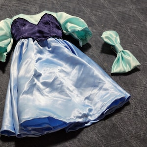 May include: A doll dress featuring a dark purple bodice with a sweetheart neckline, light blue puffed sleeves, and a matching light blue skirt. A separate light blue hair bow accessory is included with the dress, both made of a shiny satin fabric.