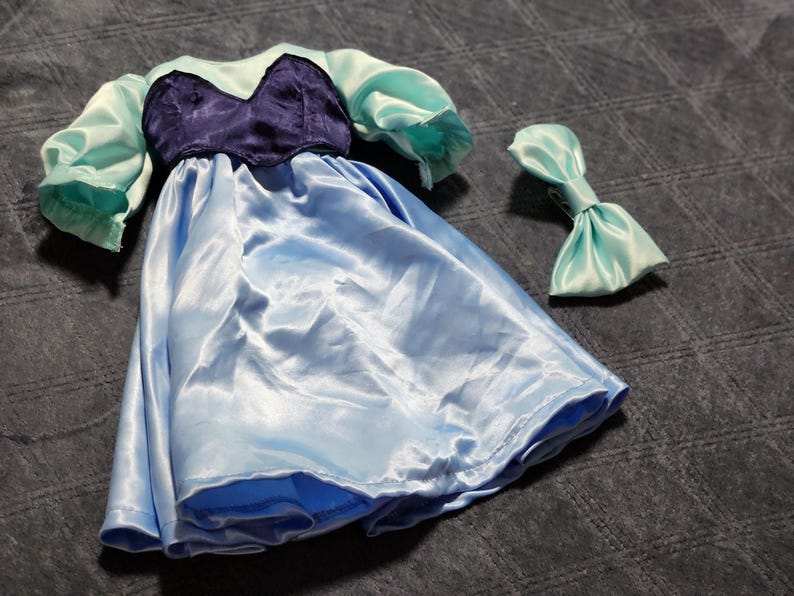 May include: A doll dress featuring a dark purple bodice with a sweetheart neckline, light blue puffed sleeves, and a matching light blue skirt. A separate light blue hair bow accessory is included with the dress, both made of a shiny satin fabric.