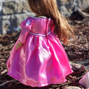 May include: A doll wearing a bright pink princess dress with long sleeves and a full skirt. The dress has white trim and a white belt. The doll has long, flowing auburn hair. The doll is standing outdoors.