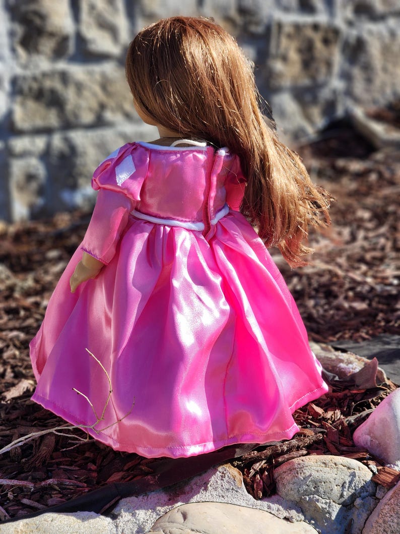May include: A doll wearing a bright pink princess dress with long sleeves and a full skirt. The dress has white trim and a white belt. The doll has long, flowing auburn hair. The doll is standing outdoors.