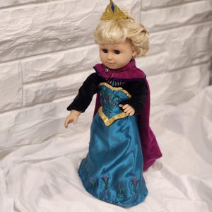 May include: A doll dressed in a teal satin gown with embroidered floral details, a black velvet top, and a purple cape. The doll has blonde hair, a gold crown, and is posed against a white background.