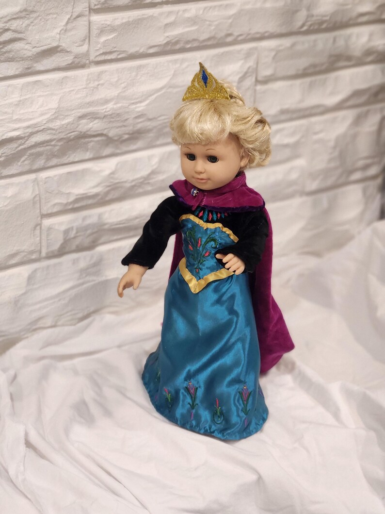 May include: A doll dressed in a teal satin gown with embroidered floral details, a black velvet top, and a purple cape. The doll has blonde hair, a gold crown, and is posed against a white background.