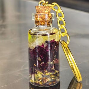 Puede incluir: Una pequeña botella de vidrio con tapón de corcho, llena de líquido transparente y flores secas. Una cadena y un llavero dorados están unidos, formando un llavero. La botella contiene elementos botánicos morados y verdes.