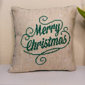 May include: A square, beige pillow with the words "Merry Christmas" embroidered in green script. The pillow has a textured appearance and a green border. It is placed on a white shelf, with a vase of white tulips to the right.