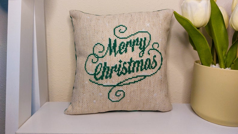 May include: A square, beige pillow with the words "Merry Christmas" embroidered in green script. The pillow has a textured appearance and a green border. It is placed on a white shelf, with a vase of white tulips to the right.