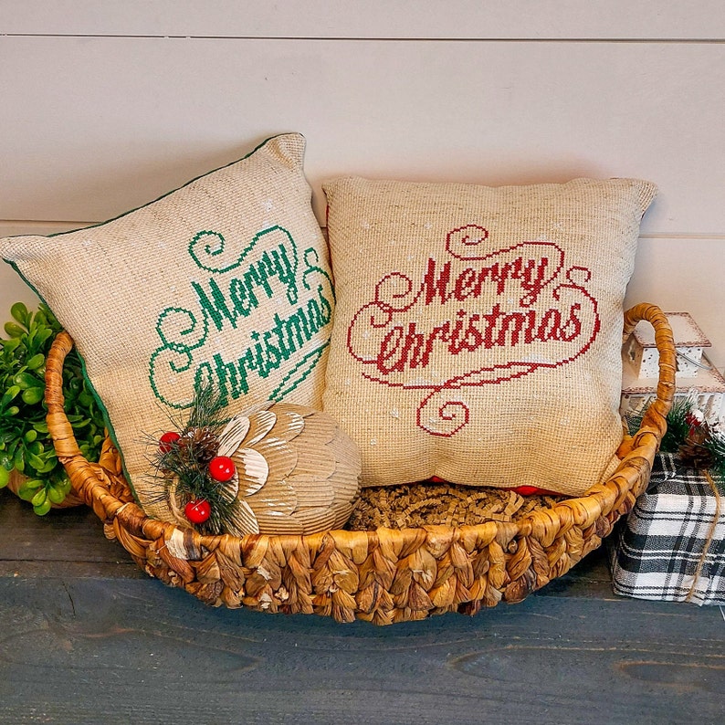 May include: Two square burlap pillows with "Merry Christmas" embroidered in green and red, nestled in a woven basket. A decorative pinecone and berry ornament sits in front of the pillows, evoking a festive holiday theme.