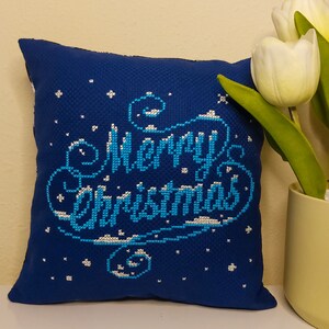 May include: A square, dark blue pillow with the words "Merry Christmas" embroidered in light blue and white. The pillow has a textured appearance and is decorated with small white star-like shapes. A vase of white tulips is to the right.