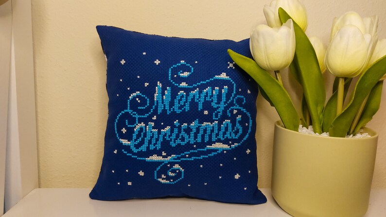 May include: A square, dark blue pillow with the words "Merry Christmas" embroidered in light blue and white. The pillow has a textured appearance and is decorated with small white star-like shapes. A vase of white tulips is to the right.