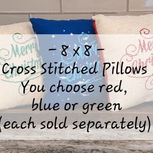 May include: Three 8x8 inch cross-stitched pillows with "Merry Christmas" text. The pillows are available in red, blue, or green. The text is cross-stitched in red, blue, and green. The pillows are on a wooden tray.