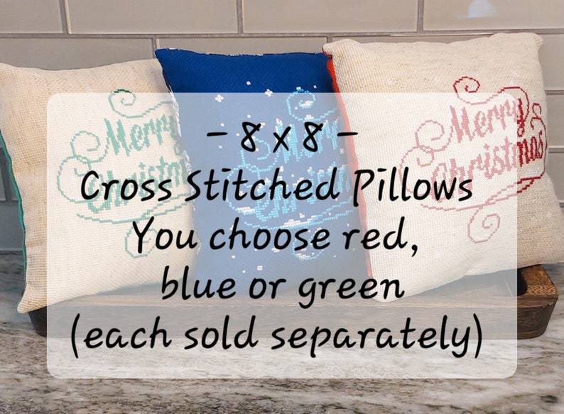 May include: Three 8x8 inch cross-stitched pillows with "Merry Christmas" text. The pillows are available in red, blue, or green. The text is cross-stitched in red, blue, and green. The pillows are on a wooden tray.