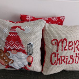 May include: Three small, square Christmas pillows. One features a gnome with a red hat and a gingerbread cookie. Another pillow has the words "Merry Christmas" in red. The third pillow is red with white snowflakes.