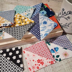 May include: A collection of square fabric coasters featuring various patterns including floral, geometric, and butterfly prints in colors such as blue, yellow, red, black, and white. The coasters are arranged in a stack and spread out on a surface.