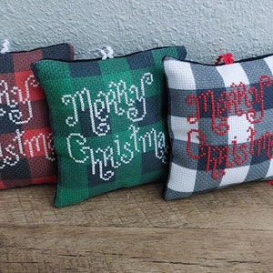May include: Three square Christmas ornaments with a plaid pattern in red, green, and gray. Each ornament has the words "Merry Christmas" embroidered in white or red thread. The ornaments are displayed on a wooden surface.