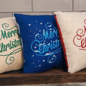 May include: Three decorative Christmas pillows with embroidered "Merry Christmas" text. One pillow is blue with white accents, and the others are off-white with green and red text. They rest on a wooden tray.