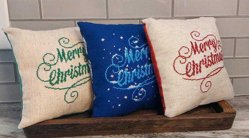 May include: Three decorative Christmas pillows with embroidered "Merry Christmas" text. One pillow is blue with white accents, and the others are off-white with green and red text. They rest on a wooden tray.