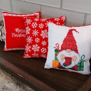 May include: Three Christmas-themed throw pillows on a wooden tray. One red pillow has white text that reads "Merry Christmas." Another red pillow features white snowflakes. The third pillow is white with a cross-stitch gnome design.