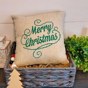 May include: A decorative pillow with the words "Merry Christmas" embroidered in green on a burlap-style fabric. The pillow is in a gray woven basket with other Christmas decorations.