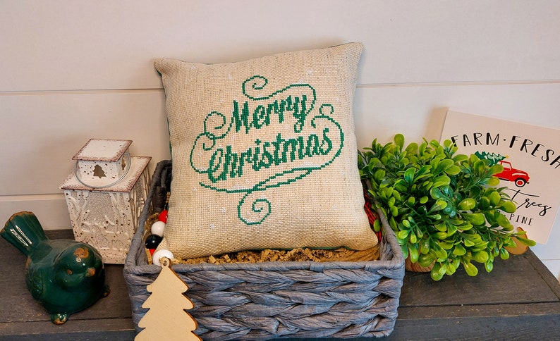 May include: A decorative pillow with the words "Merry Christmas" embroidered in green on a burlap-style fabric. The pillow is in a gray woven basket with other Christmas decorations.