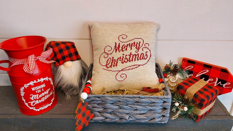 May include: A festive Christmas display featuring a red bucket with a bow, a gnome with a plaid hat, and a pillow embroidered with "Merry Christmas." A gray basket holds the pillow, and a sign that says "believe".