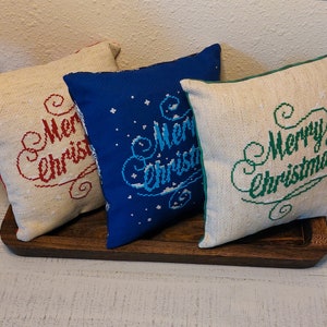 May include: Three square Christmas pillows with embroidered "Merry Christmas" text. One pillow is beige with red text, another is blue with white text, and the third is beige with green text. The pillows are on a wooden tray.