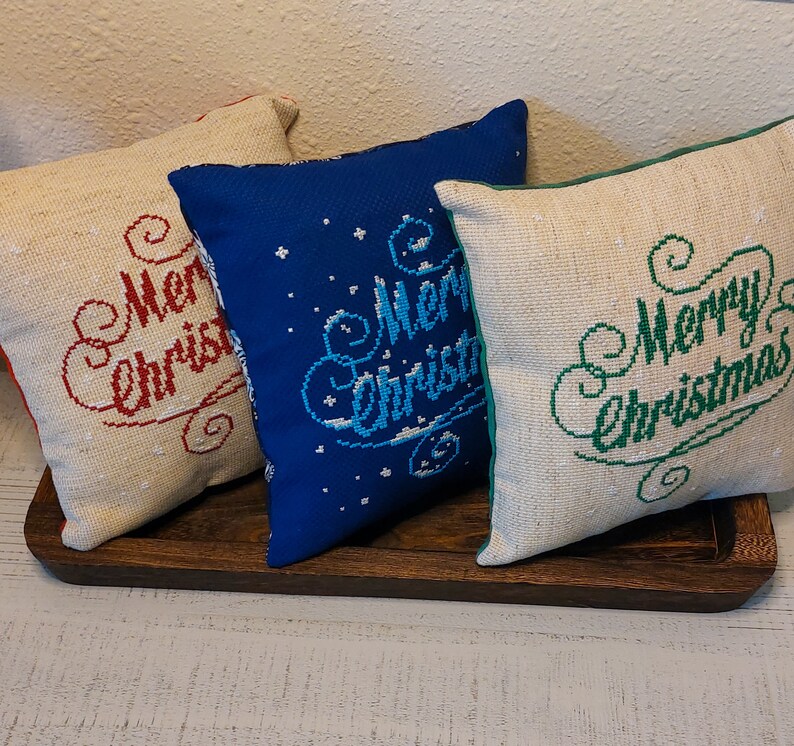 May include: Three square Christmas pillows with embroidered "Merry Christmas" text. One pillow is beige with red text, another is blue with white text, and the third is beige with green text. The pillows are on a wooden tray.