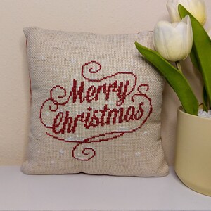May include: A square, beige Christmas pillow with the words "Merry Christmas" embroidered in red. The pillow is on a white shelf next to a vase of white tulips.