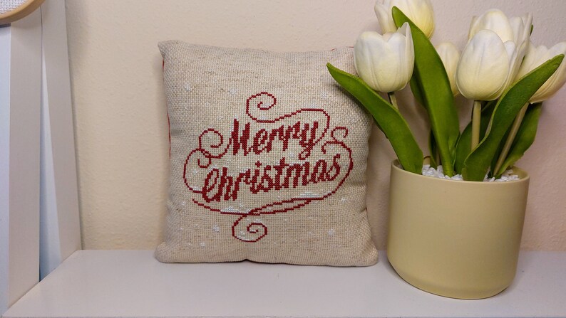 May include: A square, beige Christmas pillow with the words "Merry Christmas" embroidered in red. The pillow is on a white shelf next to a vase of white tulips.