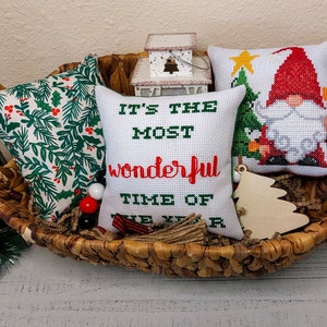 May include: A woven basket holds three decorative Christmas pillows. One pillow has a green holly pattern, another features a gnome and Christmas tree, and the third says "It's the most wonderful time of the year". A wooden Christmas tree ornament is also in the basket.