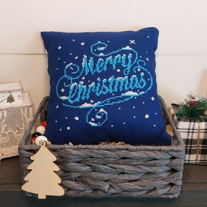 May include: A navy blue pillow with "Merry Christmas" embroidered in light blue and white, nestled in a gray woven basket. The scene includes a wooden Christmas tree ornament, a candle, and a sign that says "Farm Fresh Christmas Trees."