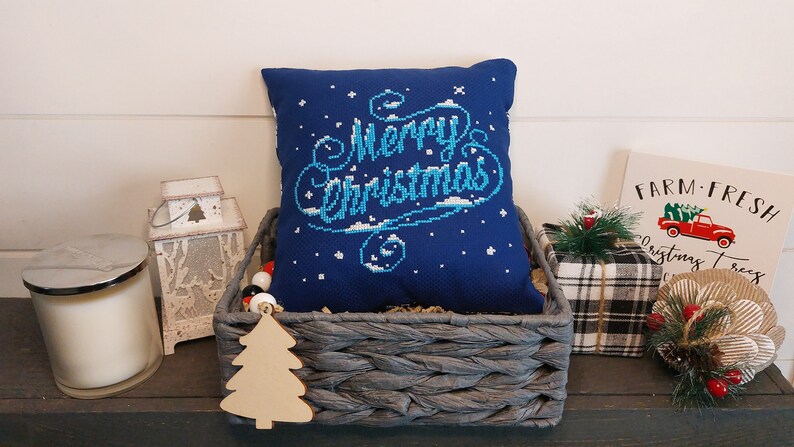 May include: A navy blue pillow with "Merry Christmas" embroidered in light blue and white, nestled in a gray woven basket. The scene includes a wooden Christmas tree ornament, a candle, and a sign that says "Farm Fresh Christmas Trees."