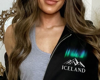 Puede incluir: Sudadera negra con cremallera y capucha, con un gráfico blanco de montañas y auroras boreales, con la palabra "ICELAND" debajo.