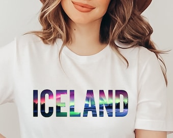 Puede incluir: Camiseta blanca con la palabra "ICELAND" impresa en un degradado de colores, que recuerda a las auroras boreales. El texto está centrado en la camiseta.