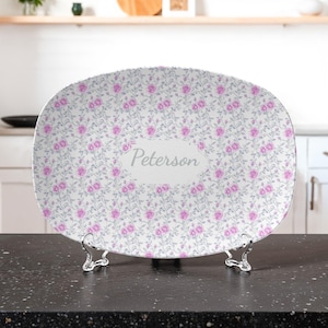 May include: Oval serving platter with a floral pattern of pink roses and gray leaves on a white background. The platter has the name "Peterson" in a light gray oval. The platter is displayed on a silver metal stand.
