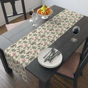 May include: A white table runner with a green and red Christmas tree pattern. The runner is laid on a dark wood table with a bowl of apples, two wine glasses, and a stack of white plates with silverware.