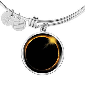 May include: A silver charm bracelet with a round charm featuring a black and gold image of a solar eclipse. The charm is attached to the bracelet with a silver ring.