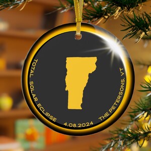 May include: A black and gold round Christmas ornament with a yellow silhouette of the state of Vermont. The ornament reads "Total Solar Eclipse 4.08.2024 The Petersons. VT"