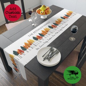 May include: A white table runner with a fall-themed design featuring colorful trees and black lines. The text "THE PETERSEN FAMILY" is printed at the top of the runner, followed by a list of names: "Pete, Jamie, Nicky, Jackson, Nan, Mike, Cindy, Mdanie, Bear".
