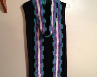May include: A crocheted scarf with white, purple, and teal stripes. The scarf is hanging on a hanger against a white wall.