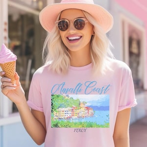 Puede incluir: Camiseta rosa claro con las palabras "Amalfi Coast" en escritura azul sobre una imagen colorida de un pueblo costero. La palabra "ITALY" está impresa debajo. La persona lleva un sombrero rosa y gafas de sol.