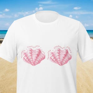 May include: White t-shirt featuring two pink seashell designs with the words "strength, confidence, love, passion, kindness, peace" in a light pink font. The shirt is made of a soft material and is perfect for a beach or casual wear.