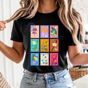 May include: Black t-shirt featuring a colorful stamp design with floral illustrations and numbers. The stamps are arranged in a grid pattern, showcasing various botanical elements. The shirt includes a postal service stamp.